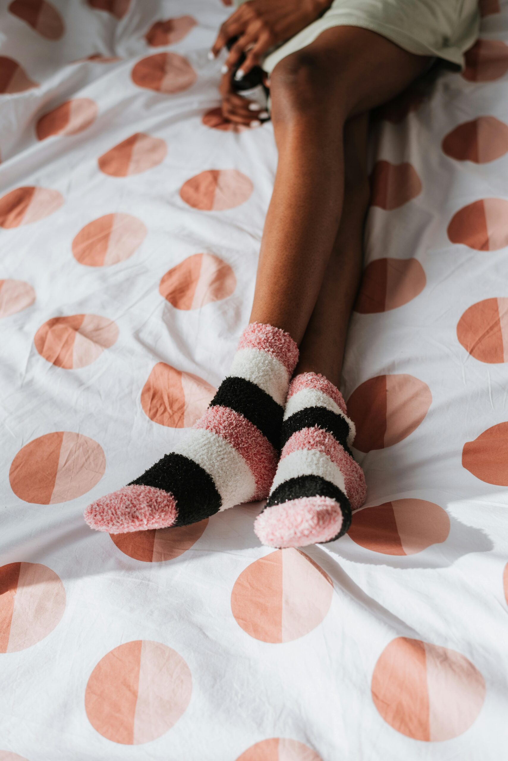 High angle of crop anonymous ethnic female in soft socks with bottle sitting with crossed legs on bed duvet in sunlight